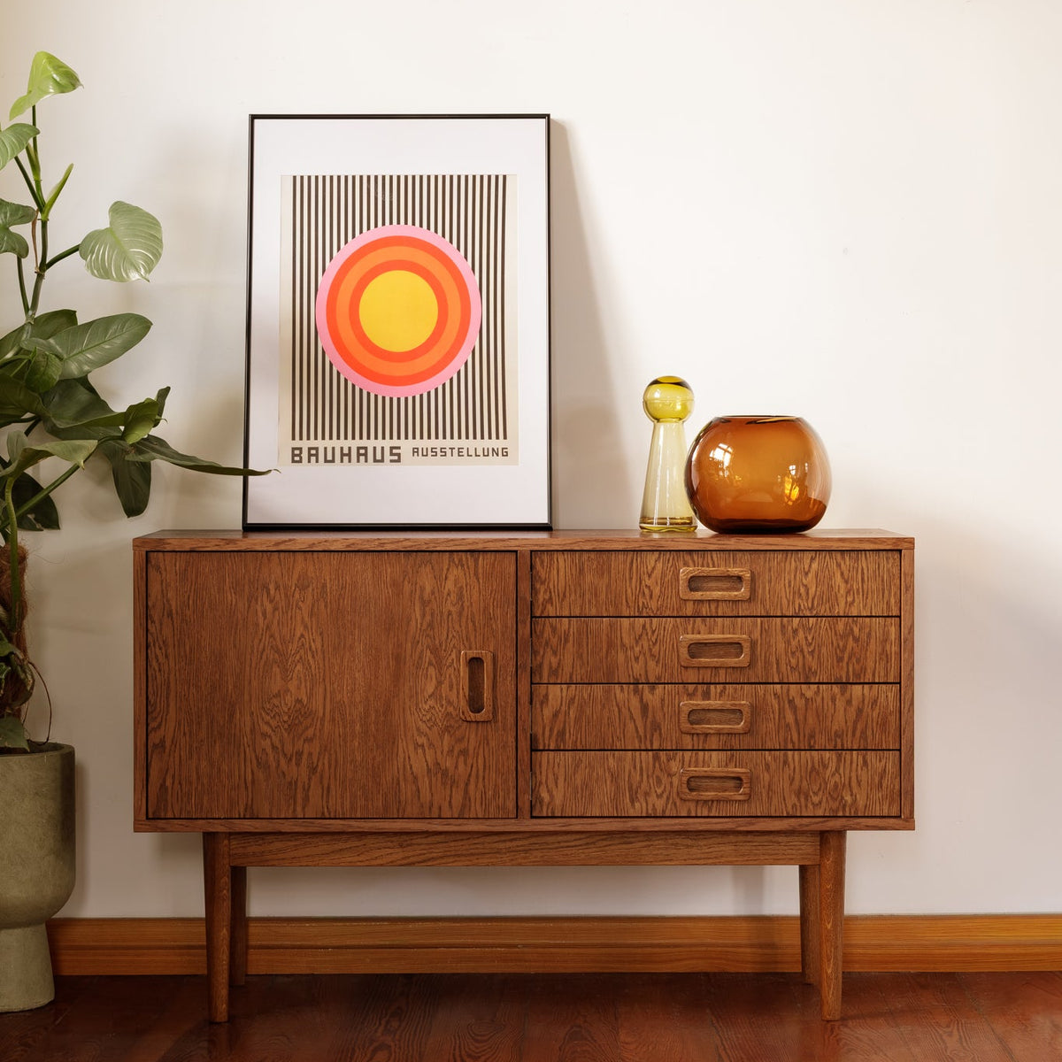 A mid-century modern wooden sideboard cabinet with a cabinet door and four drawers, styled in a room with a white wall, wooden floor, a potted plant, and framed art.