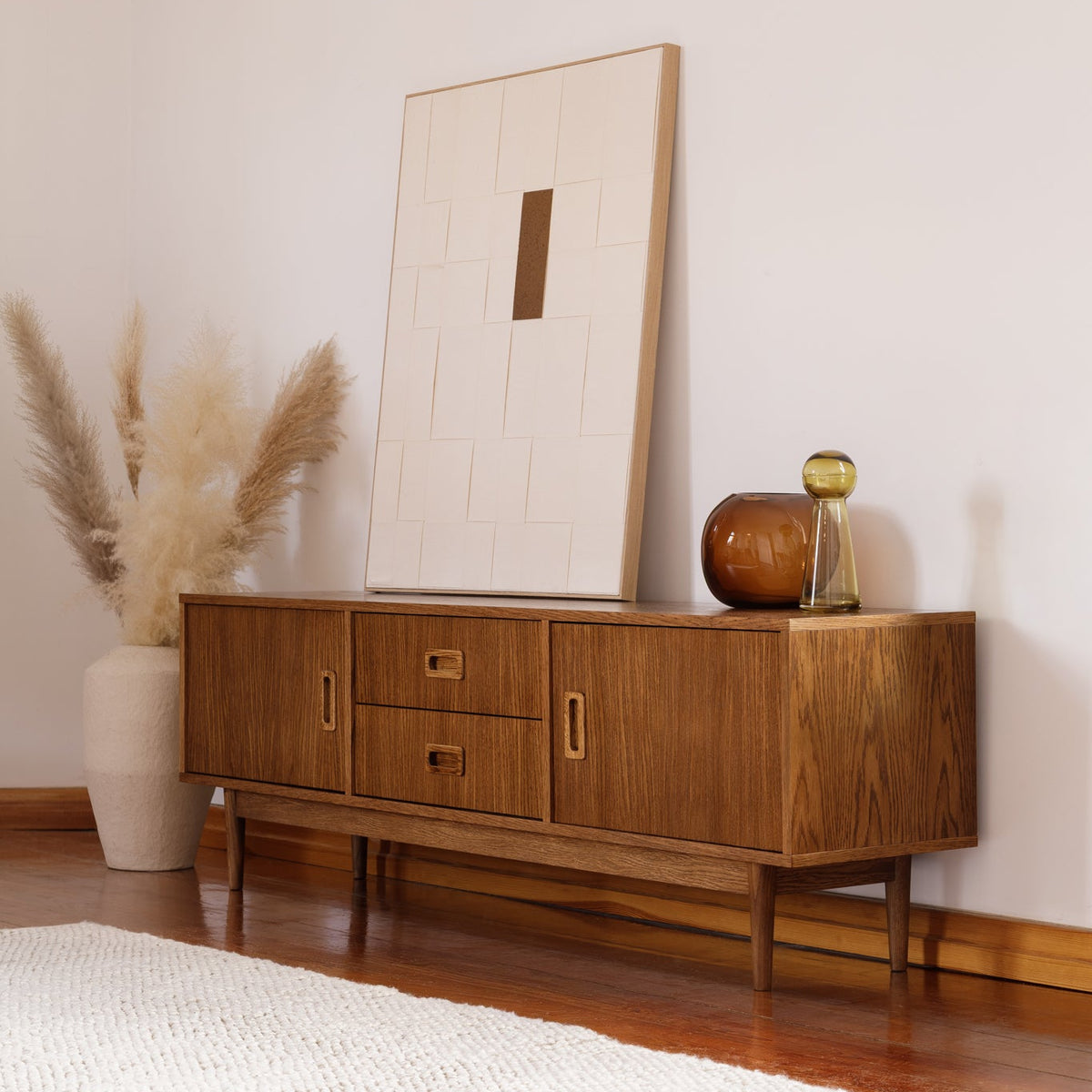 A mid-century modern wooden sideboard cabinet with three drawers and two cabinet doors, styled in a minimalist room with a large framed print, a pampas grass vase, and a small decorative object on top.