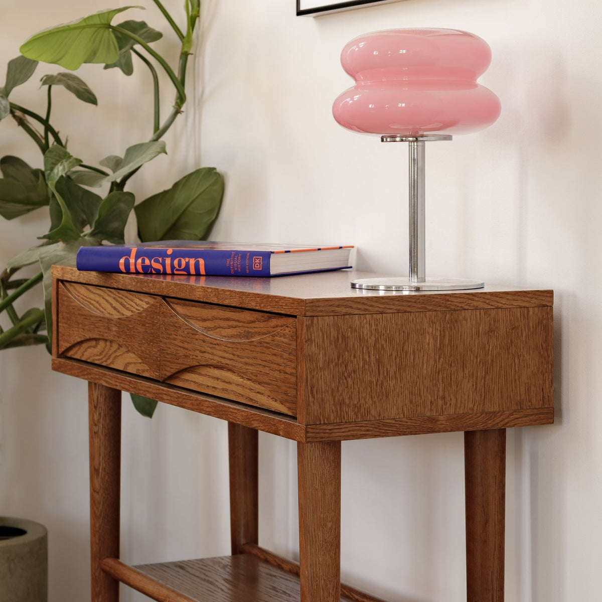 A mid-century modern wooden side table with a drawer and lower shelf, styled with a pink bubble table lamp, a blue book, and a potted green plant against a white wall.