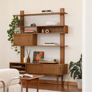 A mid-century modern wooden shelving unit and sideboard cabinet in a warm medium brown finish, styled in a living room setting with books, decorative objects, plants, a small wooden table, and a cream armchair.