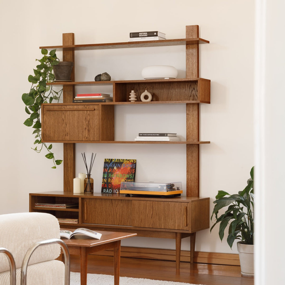 A mid-century modern wooden shelving unit and sideboard cabinet in a warm medium brown finish, styled in a living room setting with books, decorative objects, plants, a small wooden table, and a cream armchair.