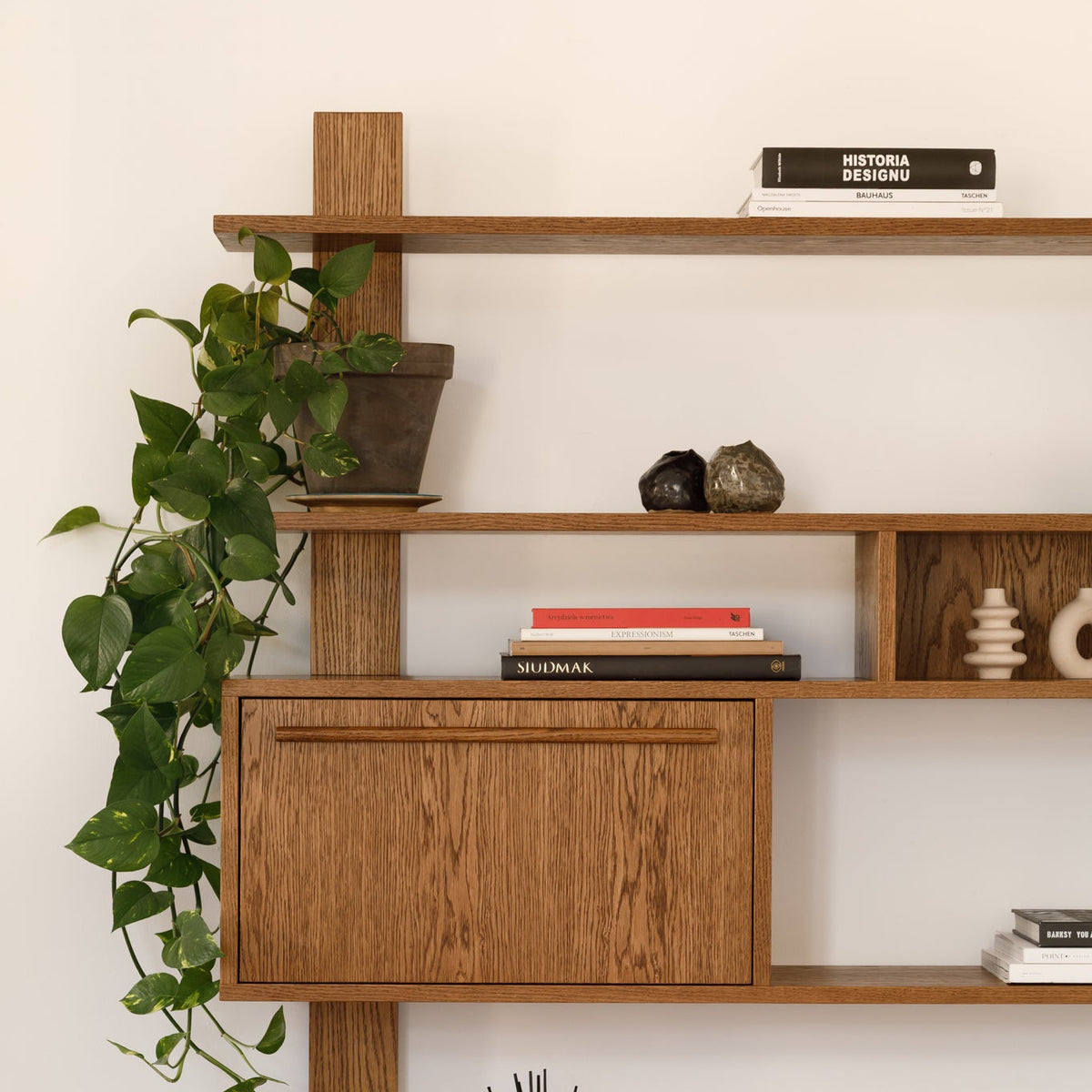 A mid-century modern wooden shelving unit with a warm medium-brown finish, styled with books, a potted trailing green plant, candles, a reed diffuser, and decorative objects against a white wall.