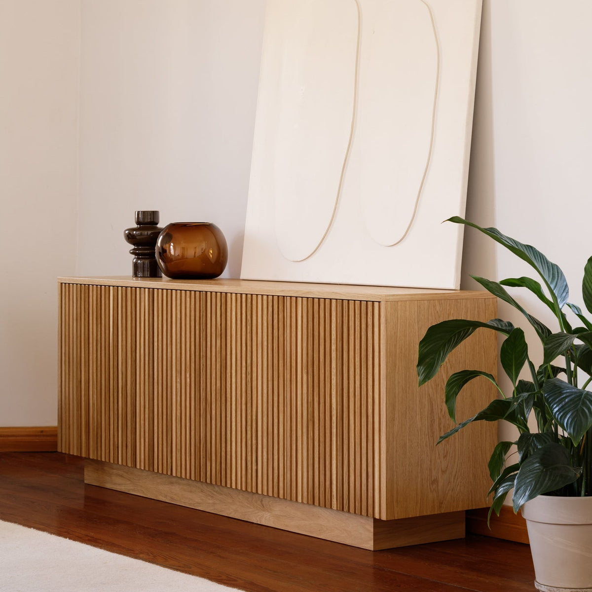 A light oak wood sideboard cabinet with vertical fluted detailing, styled in a minimalist room with a large cream abstract canvas leaning behind it, decorative objects on top, and a potted green plant on a dark wood floor.