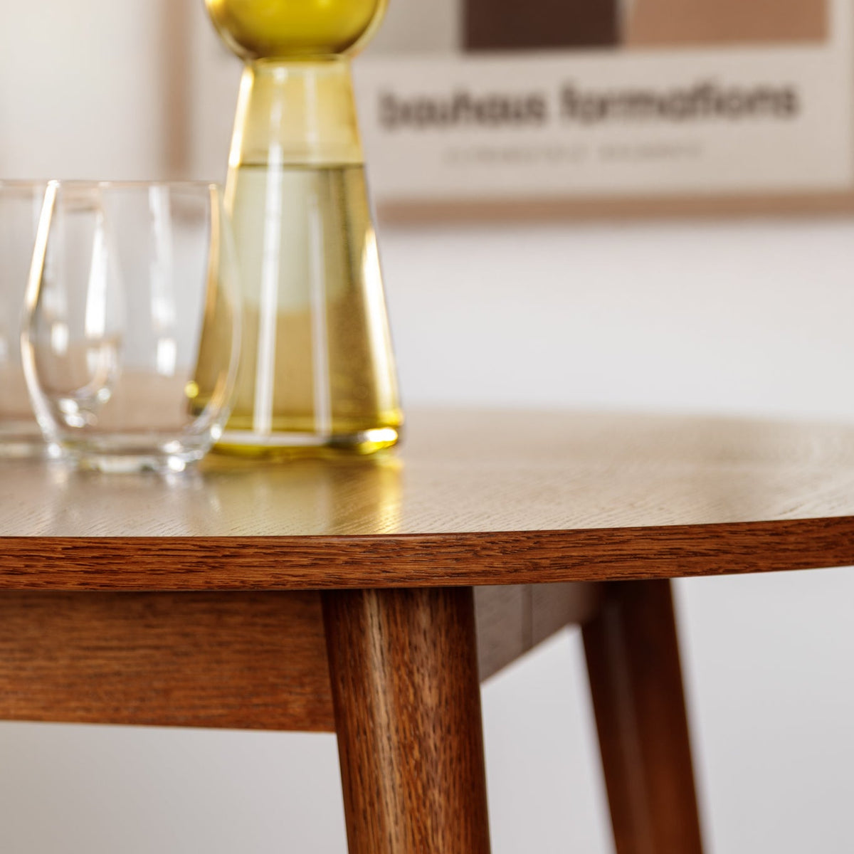 A clear glass carafe with a yellow-tinted glass stopper sits on a wooden table next to a clear drinking glass, with a modern abstract art print in the background.