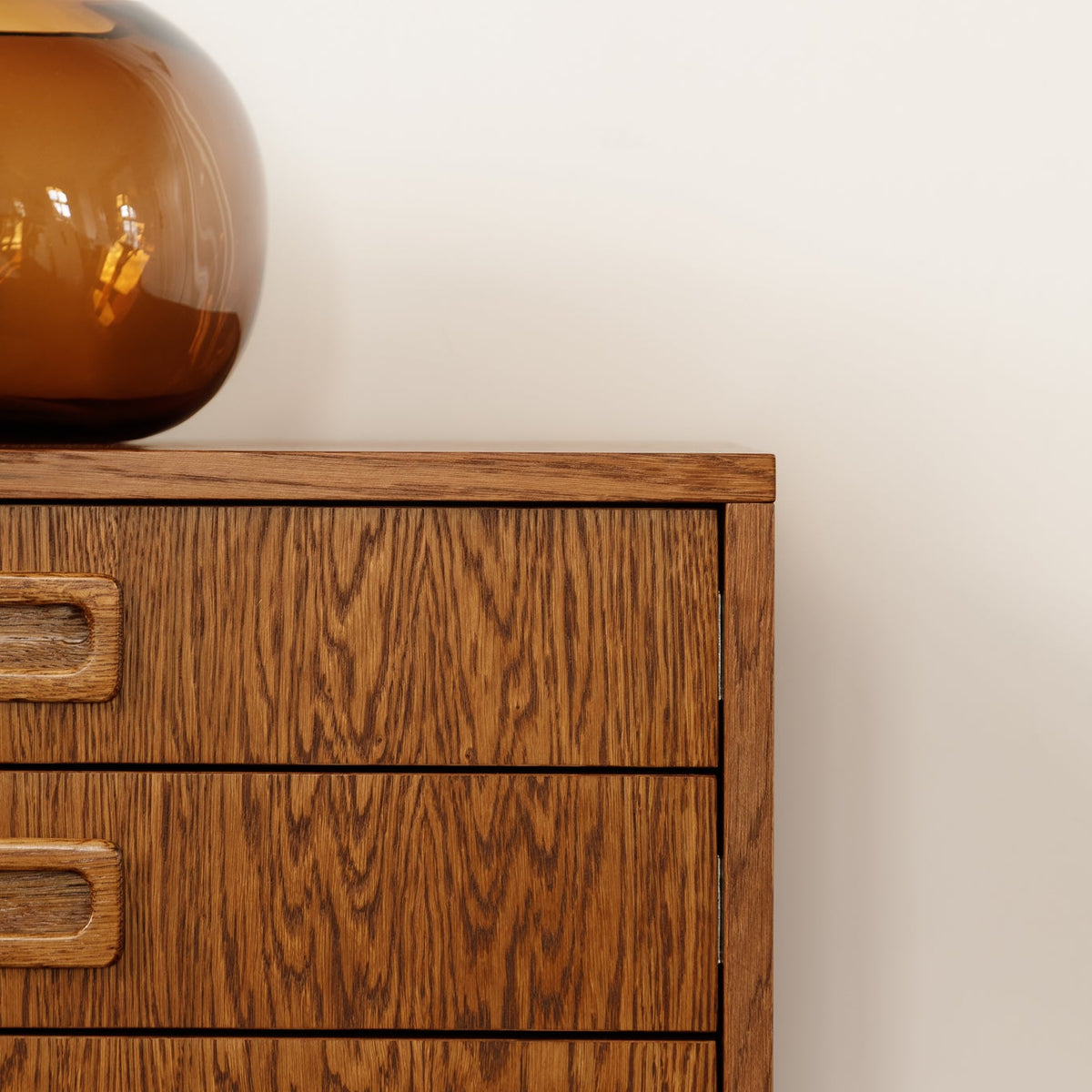 A close-up of a mid-century modern wooden chest of drawers with a warm brown oak grain finish and recessed drawer pulls, topped with an amber glass vase against a plain off-white wall.