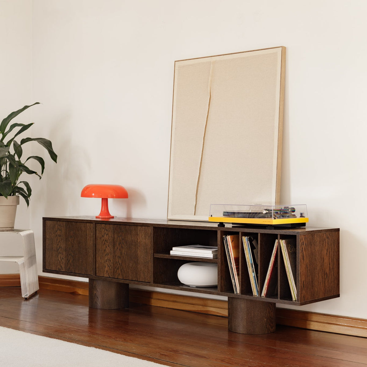 A mid-century modern wooden media console in a warm walnut finish, styled in a minimalist living room with a potted plant, orange table lamp, framed abstract art, and a yellow vinyl record player.