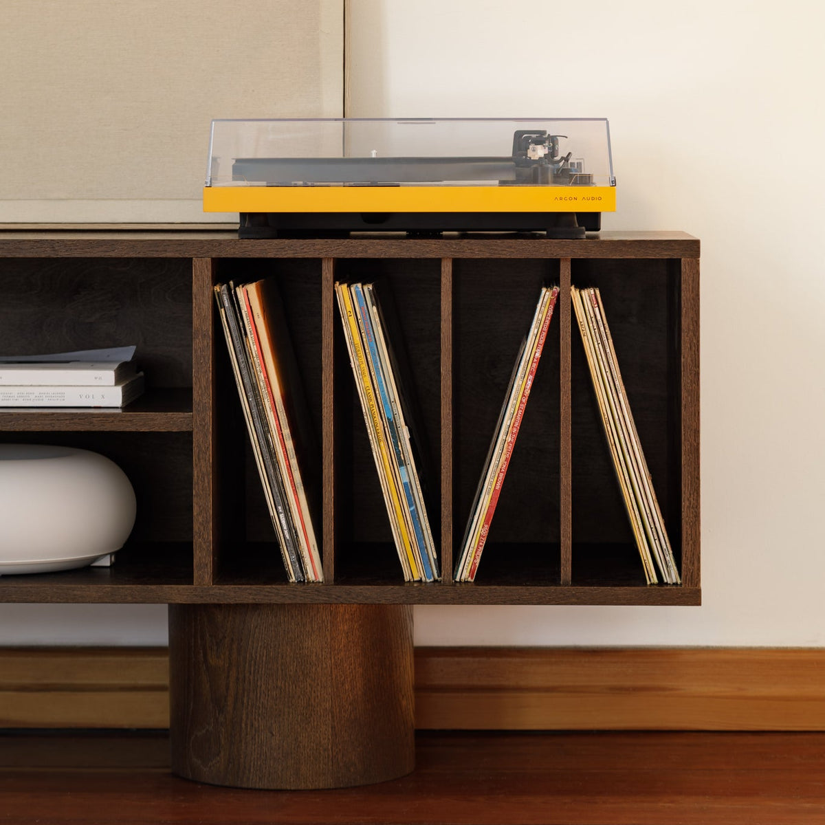 A yellow and black turntable with a clear dust cover sits on top of a dark wood floating shelf unit holding vinyl records, set against a warm beige wall and wood floor.